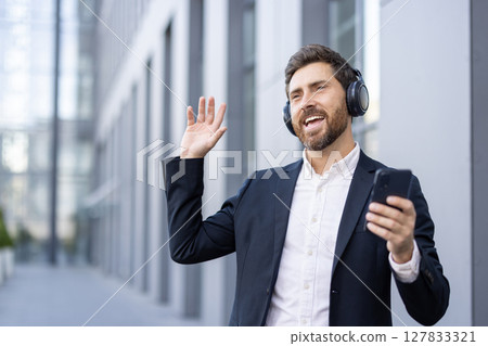 A smiling businessman in a suit waves while listening to music on headphones outside a modern building. A smiling businessman in a suit waves while listening to music on headphones outside a modern building. 127833321