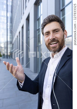 A smiling businessman is on a video call in front of a modern building, wearing a suit and using earphones, presenting with his hand 127833325