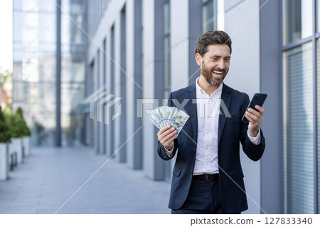 A smiling businessman in a suit holds cash and his phone, possibly celebrating a financial success outside the office. A smiling businessman in a suit holds cash and his phone, possibly celebrating a financial success outside the office. 127833340