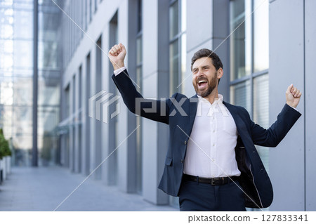 A businessman celebrates a victory, expressing joy with raised arms and an open-mouthed expression, outdoors. A businessman celebrates a victory, expressing joy with raised arms and an open-mouthed expression, outdoors. 127833341