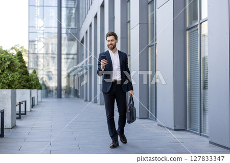A smartly dressed businessman walks outdoors while checking his phone. He is carrying a briefcase, showcasing a modern professional lifestyle near a building. A smartly dressed businessman walks outdoors while checking his phone. He is carrying a briefcase, showcasing a modern professional lifestyle near a building. 127833347