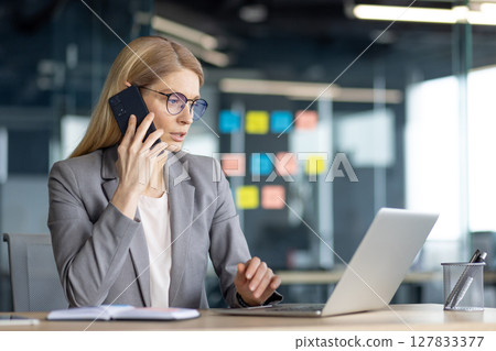 A focused businesswoman in glasses, on a phone call, works on her laptop at her desk. The office has sticky notes on the wall. A focused businesswoman in glasses, on a phone call, works on her laptop at her desk. The office has sticky notes on the wall. 127833377