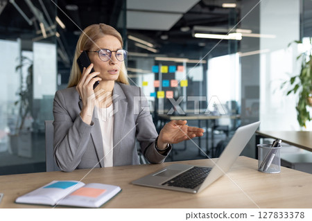Businesswoman in a grey suit is talking on the phone while gesturing with her hand as she works, at her desk with a laptop in an office. Businesswoman in a grey suit is talking on the phone while gesturing with her hand as she works, at her desk with a laptop in an office. 127833378