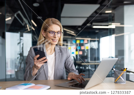 A businesswoman in glasses works on her laptop while holding her phone, in a modern office environment. She's likely multitasking. 127833387