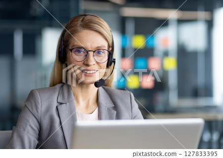 A friendly blonde woman with glasses wears a headset, smiling at the camera while working on a laptop in an office setting. 127833395