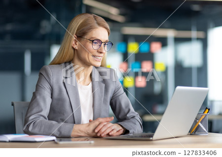 A smiling businesswoman in glasses works on a laptop at her desk in a modern office setting, looking positive. 127833406