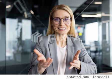 Businesswoman smiling and gesturing talking looking at camera, woman working in office using laptop for online video call, remote conference meeting. 127833411