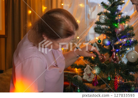 Woman decorates Christmas tree with ornaments in cozy living room during festive holiday season 127833593