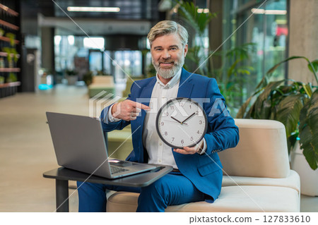 Mature businessman with laptop in lobby showing time on wall clock and pointing the finger at camera Mature businessman with laptop in lobby showing time on wall clock and pointing the finger at camera 127833610