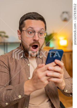 Caucasian young man guy using smartphone shocked by sudden victory sitting in home office room Caucasian young man guy using smartphone shocked by sudden victory sitting in home office room 127833612