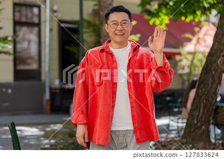 Asian man smiling friendly at camera, waving hands hello, hi, greeting or goodbye in city street Asian man smiling friendly at camera, waving hands hello, hi, greeting or goodbye in city street 127833624
