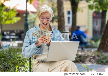 Happy rich young woman using laptop winner victory holding cash money in dollar banknotes in city Happy rich young woman using laptop winner victory holding cash money in dollar banknotes in city 127833626