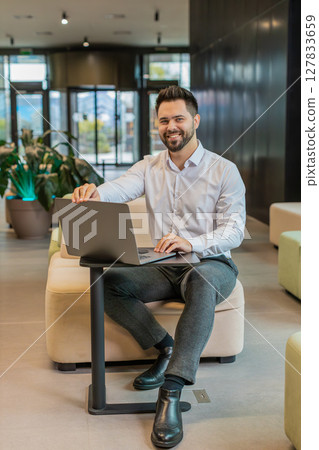 Young Caucasian businessman in while shirt opens laptop and starts working sitting in office lobby Young Caucasian businessman in while shirt opens laptop and starts working sitting in office lobby 127833659
