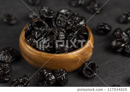 old bowl filled with dried prunes made from small plums, dark-colored dried plum fruits poured into a wooden bowl, closeup old bowl filled with dried prunes made from small plums, dark-colored dried plum fruits poured into a wooden bowl, closeup 127834829