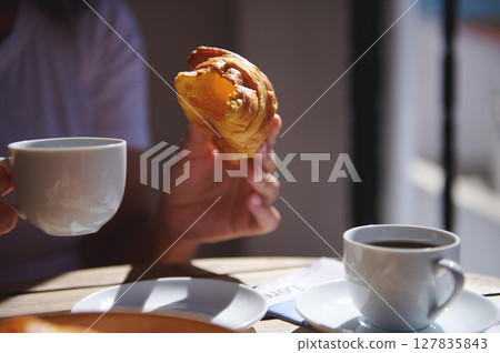 Close-up of a person enjoying a coffee and croissant outdoors. Warm sunlight creates a cozy atmosphere, highlighting the morning ritual of coffee paired with baked delights. 127835843