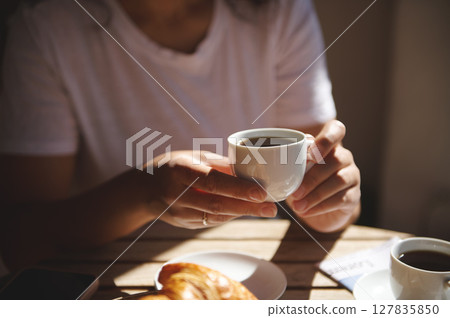 Close-up of hands holding a cup of coffee with a croissant on the table. A cozy moment enjoying breakfast in a bright and warm setting. 127835850