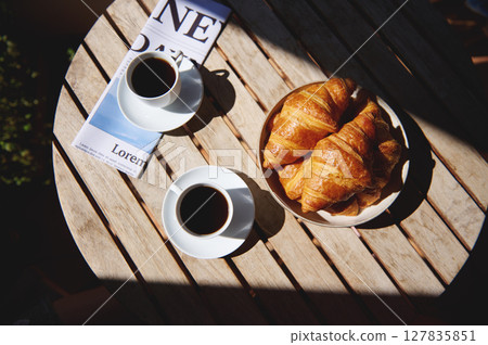 Image of a wooden table with cups of coffee, crispy croissants, and a newspaper, creating a tranquil breakfast atmosphere in soft morning light. 127835851