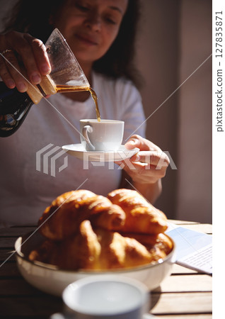 A scene of breakfast preparation as coffee is poured into a cup, accompanied by golden, flaky croissants on a wooden surface, evoking notions of comfort, warmth, and culinary delight. 127835871