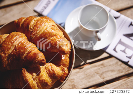 Fresh golden croissants served with a cup of coffee and a newspaper on a rustic wooden table, portraying a cozy breakfast. Fresh golden croissants served with a cup of coffee and a newspaper on a rustic wooden table, portraying a cozy breakfast. 127835872