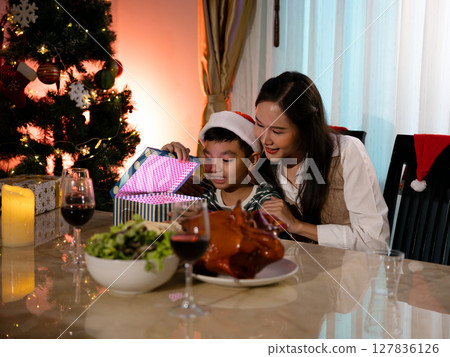 Excited child opening glowing Christmas gift box with smiling mother beside festive dinner table and decorated tree Excited child opening glowing Christmas gift box with smiling mother beside festive dinner table and decorated tree 127836126