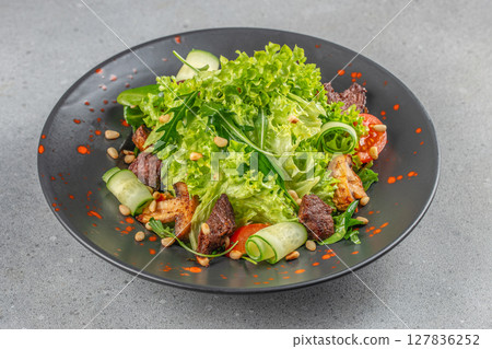 Fresh green salad with grilled meat, cucumber rolls, cherry tomatoes, and toasted pine nuts served on a black plate with artistic red sauce splashes 127836252