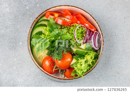 A fresh and colorful vegetable salad with cucumbers, tomatoes, red onions, bell peppers, lettuce, and herbs arranged in a rustic bowl on a gray stone surface 127836265
