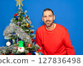 Bearded Hispanic man in his 40s wearing a red sweater posing satisfied and happy next to a Christmas tree, isolated on a blue studio background. Christmas concept. 127836498