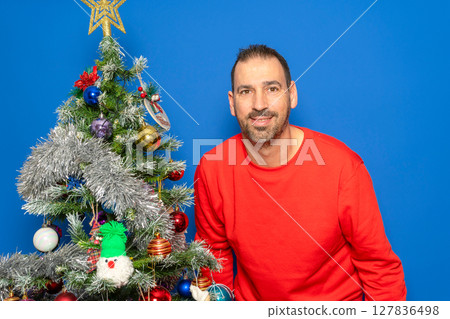 Bearded Hispanic man in his 40s wearing a red sweater posing satisfied and happy next to a Christmas tree, isolated on a blue studio background. Christmas concept. Bearded Hispanic man in his 40s wearing a red sweater posing satisfied and happy next to a Christmas tree, isolated on a blue studio background. Christmas concept. 127836498