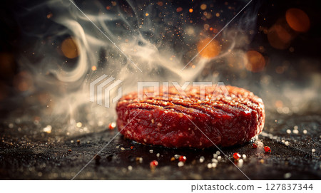 Sizzling red beef hamburger patty emitting smoke on a dark grill surface 127837344