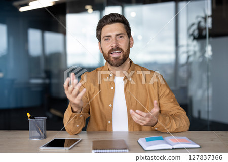 A man in a video call gestures enthusiastically while talking, likely at a desk in an office setting. A man in a video call gestures enthusiastically while talking, likely at a desk in an office setting. 127837366