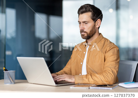A focused businessman working on a laptop in an office environment, showcasing concentration and productivity. A focused businessman working on a laptop in an office environment, showcasing concentration and productivity. 127837370