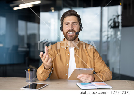 Confident businessman using headset, engaging in virtual meeting. Enthusiastic expression, holding notebook, gesturing during online discussion. Tablet and phone on desk enhance modern office setting 127837373