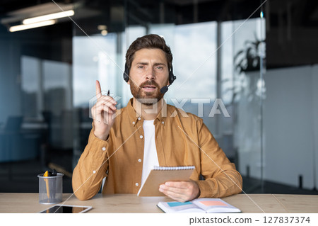 Businessman with headset taking notes during virtual meeting, emphasizing communication and leadership in office setting. Person making a point, highlighting professional presentation. 127837374