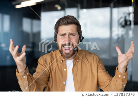 Businessman wearing headset looking frustrated handling a difficult customer support call. Professional man displaying feelings in tech office setting. Concept service communication. Businessman wearing headset looking frustrated handling a difficult customer support call. Professional man displaying feelings in tech office setting. Concept service communication. 127837376