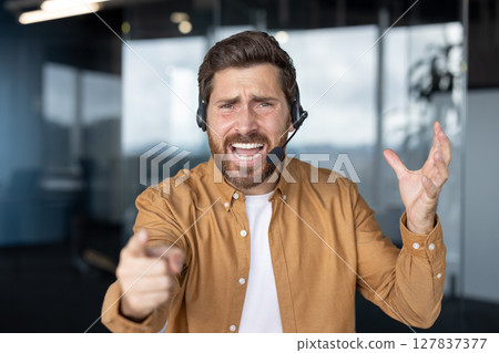 Frustrated customer service representative wearing headset in office expressing dissatisfaction through gestures. Pointing forward with annoyed facial expression suggests stress in workplace. 127837377
