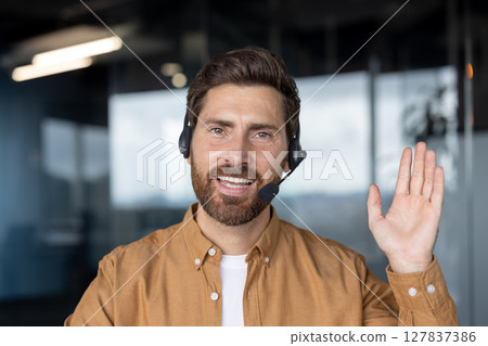 Confident man in office wears headset, smiling warmly while waving. Represents customer service, communication skills, professionalism. Ideal for business, support, tech, positive work environment. Confident man in office wears headset, smiling warmly while waving. Represents customer service, communication skills, professionalism. Ideal for business, support, tech, positive work environment. 127837386