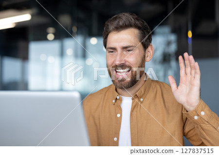 Man in brown shirt smiling during video call, greeting with raised hand in modern office space. Engaged in friendly online communication using laptop, conveying warmth and connection. 127837515