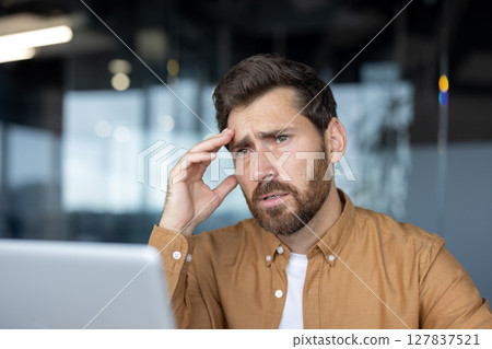 Mature businessman feeling stressed while looking at laptop, sitting in modern office. Expressive portrait captures stress and concentration. Ideal for business, stress, work themes. Mature businessman feeling stressed while looking at laptop, sitting in modern office. Expressive portrait captures stress and concentration. Ideal for business, stress, work themes. 127837521
