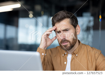 Man in office setting deeply focused on laptop, looking thoughtful, indicating problem-solving. Office environment suggests professional setting with colleagues and conversations unfolding. Man in office setting deeply focused on laptop, looking thoughtful, indicating problem-solving. Office environment suggests professional setting with colleagues and conversations unfolding. 127837523