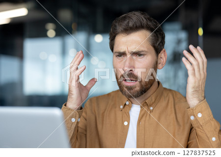 Mature businessman showing frustration in modern office, hands raised in disbelief. Professional expressing stress, likely due to unexpected challenge during important work task on computer. 127837525