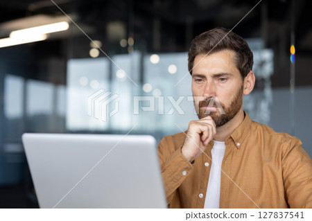 Mature businessman working on laptop in modern office, deep in thought. Man dressed in casual attire displays concentration and focus, reflecting on business strategies and solutions. 127837541
