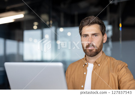 Mature businessman with a confident expression using laptop in modern office. He exudes professionalism, focus, determination, dressed in casual attire, working on business tasks. 127837543