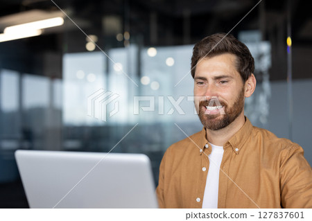 Confident business man smiling while working on laptop in modern office setting. Professional appearance. Reflects positivity, focus, success in work. Ambiance inspires productivity. Confident business man smiling while working on laptop in modern office setting. Professional appearance. Reflects positivity, focus, success in work. Ambiance inspires productivity. 127837601