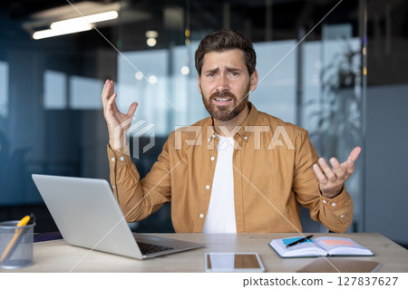 Frustrated businessman gestures at desk with laptop, notebook, phone, suggesting tech issue or confusion in office. Uncertainty and stress in workplace captured in moment. 127837627