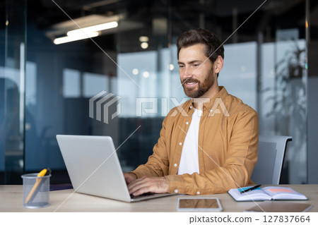 Mature businessman in modern office focused on laptop. Smiling, attentive, works with notebook and smartphone on desk. Represents productivity, concentration, professional success. 127837664