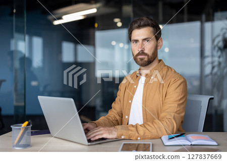 Confident mature businessman using laptop in modern office setting. Open notebook, pen, smartphone on desk indicate organized, productive work environment. Business, technology, focus. Confident mature businessman using laptop in modern office setting. Open notebook, pen, smartphone on desk indicate organized, productive work environment. Business, technology, focus. 127837669