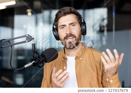 A man wearing headphones speaks into a microphone in a podcast studio setting, gesturing with his hands. A man wearing headphones speaks into a microphone in a podcast studio setting, gesturing with his hands. 127837679