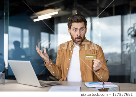 A man sits at a desk, looking shocked while holding a credit card near a laptop, possibly experiencing financial issues. A man sits at a desk, looking shocked while holding a credit card near a laptop, possibly experiencing financial issues. 127837691