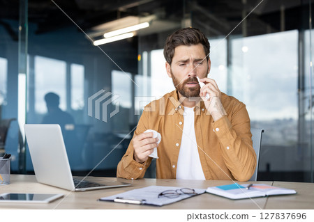 A man at his office desk is experiencing a cold, using a tissue to blow his nose while working. A man at his office desk is experiencing a cold, using a tissue to blow his nose while working. 127837696