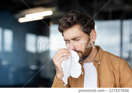 A man with a beard blowing his nose with a tissue, possibly suffering from allergies or a cold. A man with a beard blowing his nose with a tissue, possibly suffering from allergies or a cold. 127837698